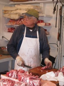 Frank "Scotty" Rees poses for a photo in his meat processing cooler. Scotty is an older white male with white hair and clear round glasses. He wears a gray shirt, denim overalls, with a green ball cap. A white apron with an R protects his clothing from the butchering process, a piece of meat in his right hand and a knife in his left. Behind him is pork drying on a shelf, the foreground of the photo features slices of pork.