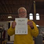 Bob Patrick is photographed with his TAAP certificate. He is an older white man with a fully white beard and hair. He wears thin wired glasses and a yellow sweater.