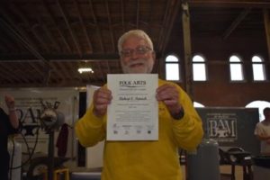 Bob Patrick is photographed with his TAAP certificate. He is an older white man with a fully white beard and hair. He wears thin wired glasses and a yellow sweater.