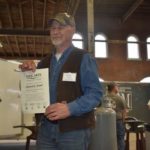 Bernard Tappel is photographed with his TAAP certificate. Tappel is an older white male with a white beard, he wears an army camoflauge baseball cap. Tappel wears a white undershirt and denim button down shirt under a brown sleeveless vest. He wears denim jeans.