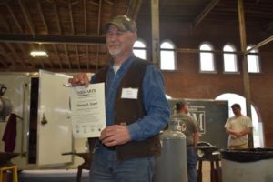 Bernard Tappel is photographed with his TAAP certificate. Tappel is an older white male with a white beard, he wears an army camoflauge baseball cap. Tappel wears a white undershirt and denim button down shirt under a brown sleeveless vest. He wears denim jeans.