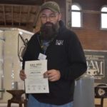 John Kirby poses for photo with his TAAP certificate extended in front of him. Kirby is a white man with long brown mustache and beard. He has dark brown eyebrows and wears thin wired glasses. Kirby wears a blue zipped up jacket, a pair of denim jeans, and a brown baseball cap.