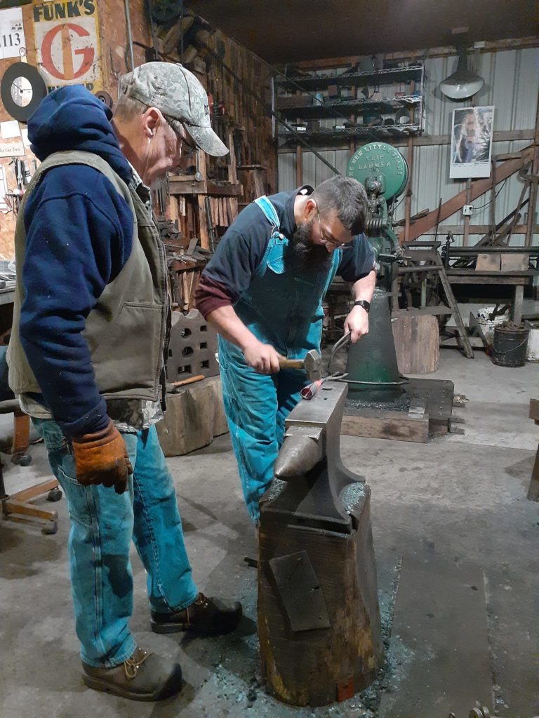 One blacksmith works at the anvil with hammer while the second observes.