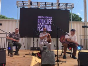 Three musicians pose on festival stage with mics and monitors. and Smithsonian Folklife Festival banner behind them.