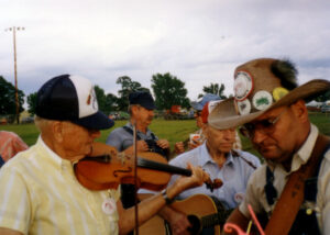 Man on left in cap playing fiddle outsisde with guitarist in straw cowboy hat on right, outside with other musicians in background. 