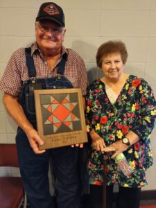 A man in a tractor cap, plaid shirt and overalls holds a red, gray, and black plaque standing next to a woman with short red curly hair wearing a floral tunic.