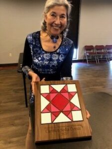 Woman with shoulder length gray hair in blue dress holds plaque with red, pink, and white quilt pattern