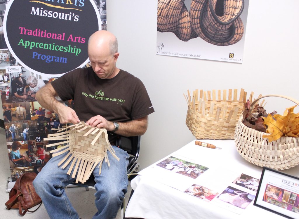 Man in brown tshirt and jeans weaves wooden splints to start a basket. 