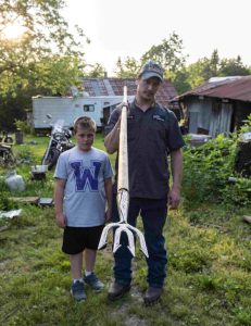 Boy in tshirt and shorts stands outside next to father in cap and uniform who is holding a four-pronged gig mounted to a long woodenpole.