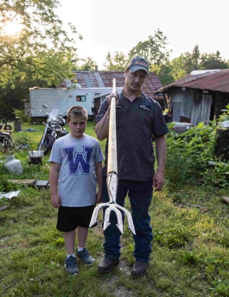 Boy in t-shirt and shorts stands outside next to father who is holding a four-pronged gig mounted to a long pole.