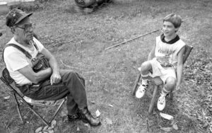 Older man in cap and overalls and young boy sit on folding chairs outside in black and white photo from the 1990s. 