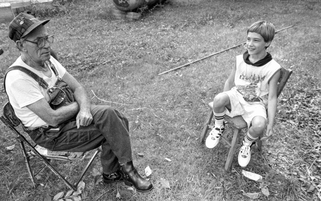 Older man and young boy sit on folding chairs outside in black and white photo from the 1990s.