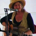 Seated woman in straw hat talks into microphone while holding her guitar.