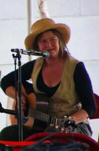 Seated woman in straw hat talks into microphone while holding her guitar. 