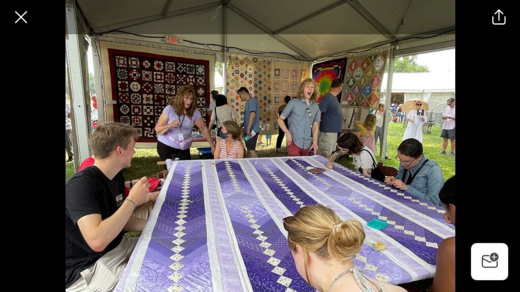 People surround a purple and white striped quilt on a frame. 