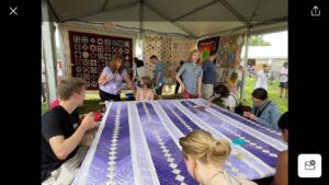 People surround a purple and white striped quilt on a frame.