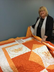 Woman with blond hair, scarrf, and black suit holds orange quilt for display.