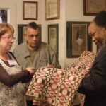 An African American man in black robe holds a green, white, and pink hand-woven coverlet while an African American man in army green jacket and white woman with glasses look on.
