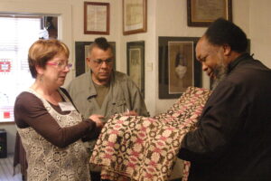 An African American man in black robe holds a green, white, and pink hand-woven coverlet while an African American man in army green jacket and white woman with glasses look on.