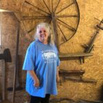 Women with long gray hair and blue t-shirt smiles in front of a spinning wheel display.