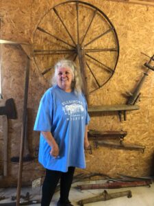 Women with long gray hair and blue t-shirt smiles in front of a spinning wheel display.