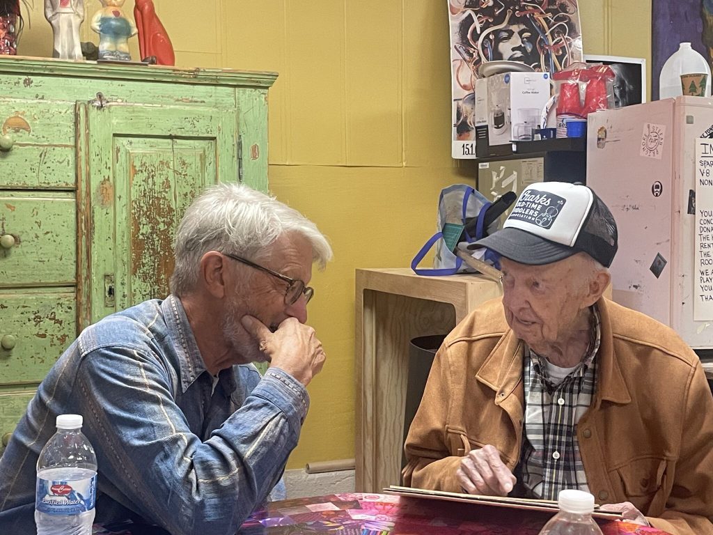 Seated at a table, speaker Barry Bergey on the right listens as Gordon McCann tells a story about documenting folk arts in the past.