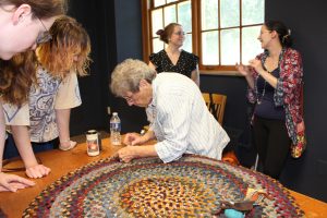 Women stand around a table as one of them shows how to stitch a rug. 